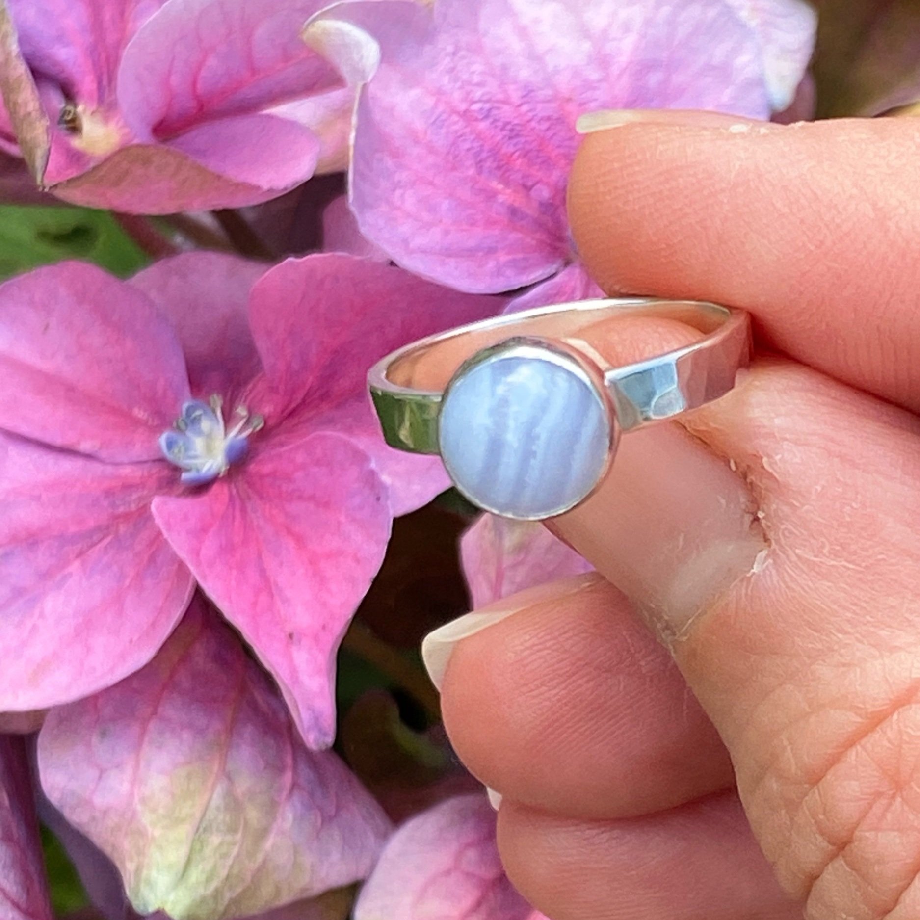 Walter and Nancy Jewellery's handmade, sterling silver and blue lace agate ring shown being held in front of some hydrangea.