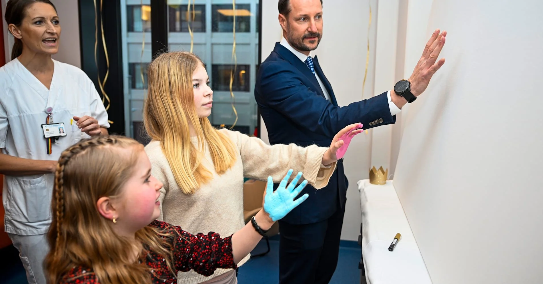 To unge jenter og en voksen mann deltar i en aktivitet, med en kvinne i sykepleieruniform som observerer. Jentene har maling på hendene, og mannen skriver på en hvit tavle.