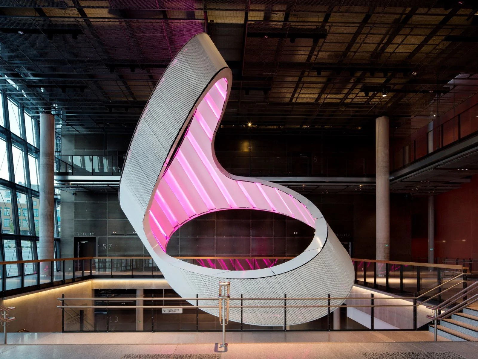Innendørs moderne arkitektonisk struktur med lysende rosa og hvite lys, omgitt av glassvegger og stålstolper.