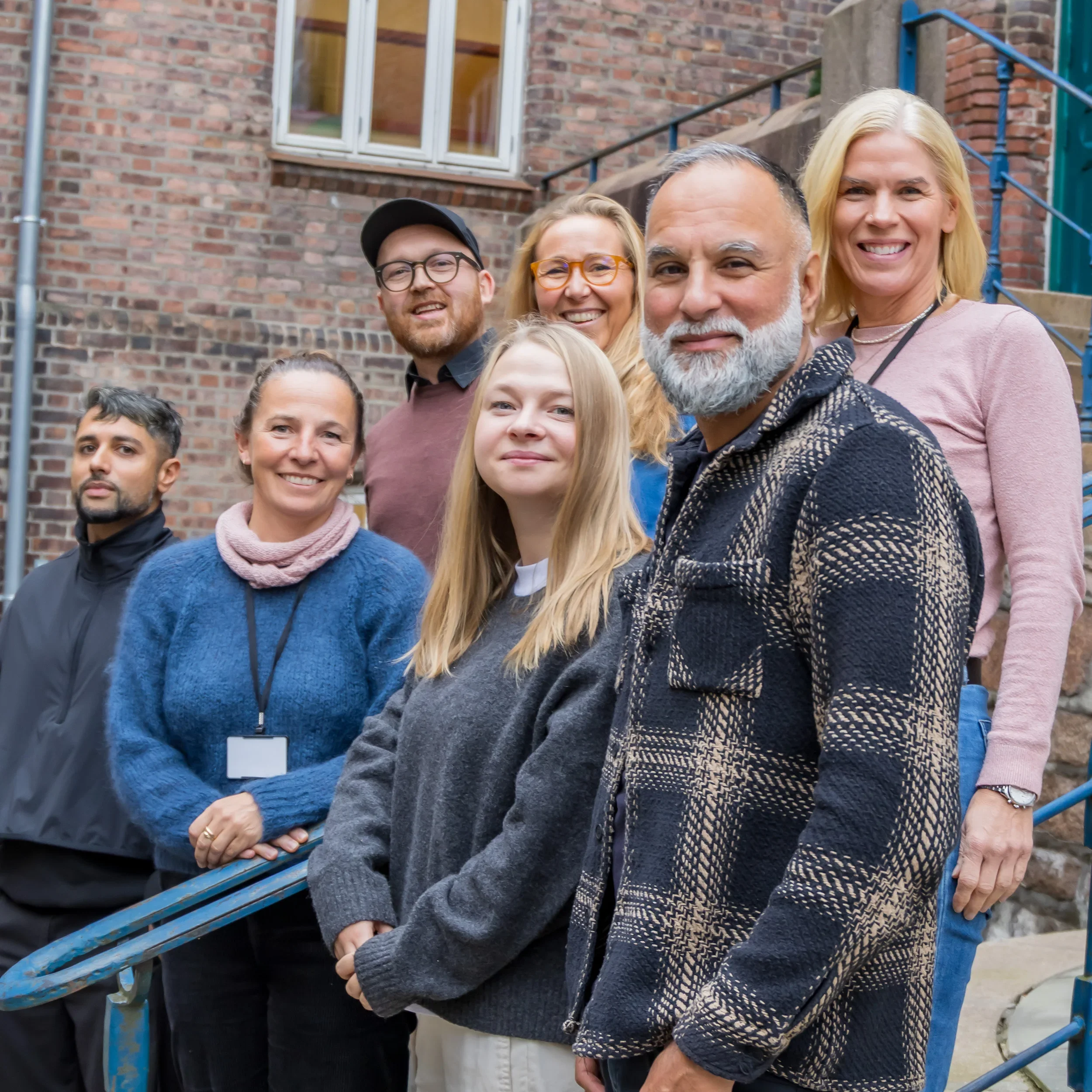 En gruppe med syv mennesker som står på en trapp foran en rød vegg, smilende og poserer for et bilde.