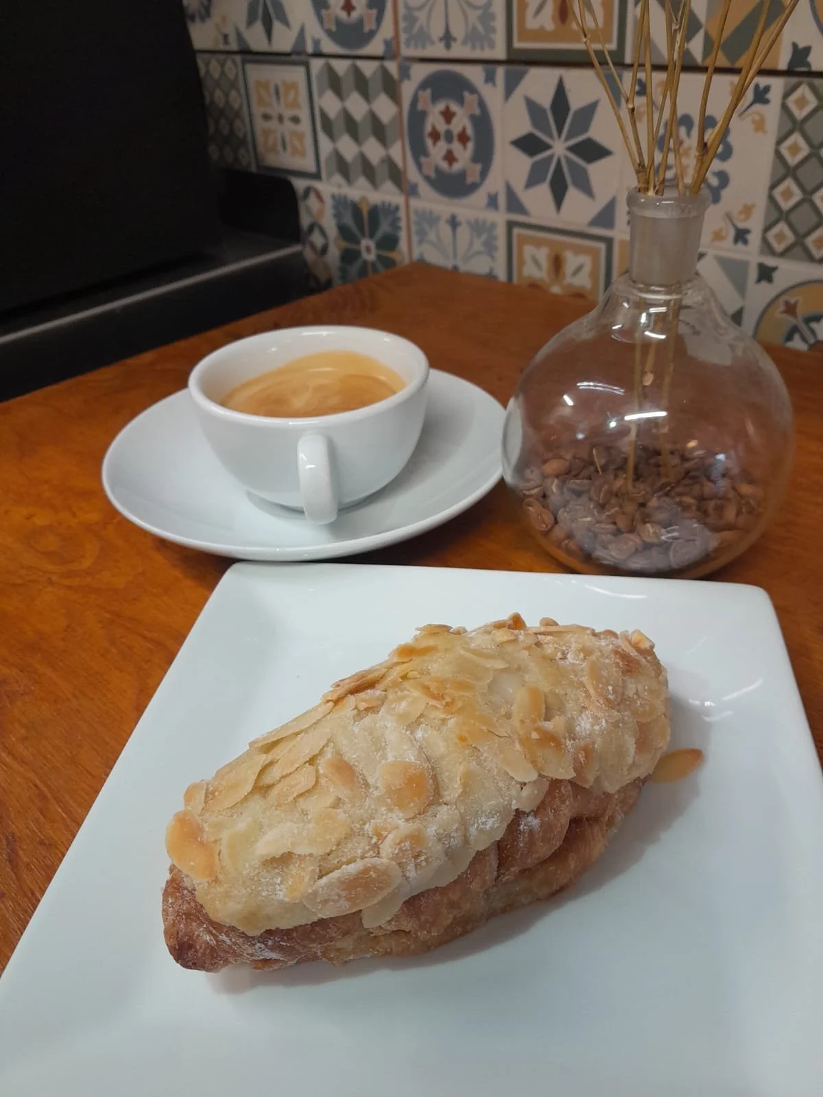 A pastry on a white plate with a cup of coffee and a glass jar with dried sticks and coffee beans on a wooden table, with decorative blue and white tiles in the background.