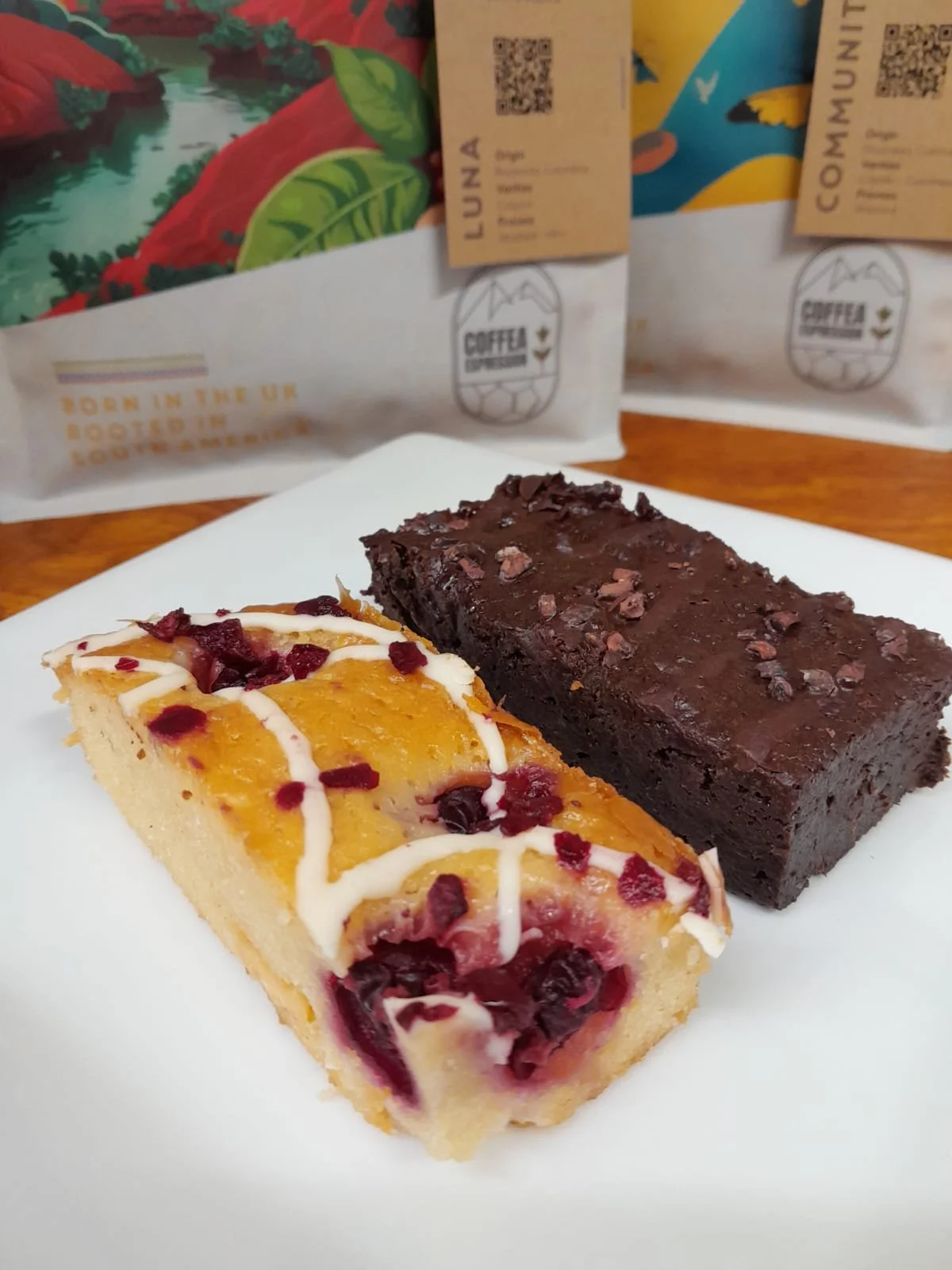 Two slices of dessert, one appears to be a cherry or berry cheesecake with white icing drizzle and dried fruit topping, the other a chocolate brownie with chocolate chunks, placed on a white plate with packaged coffee bags in the background.