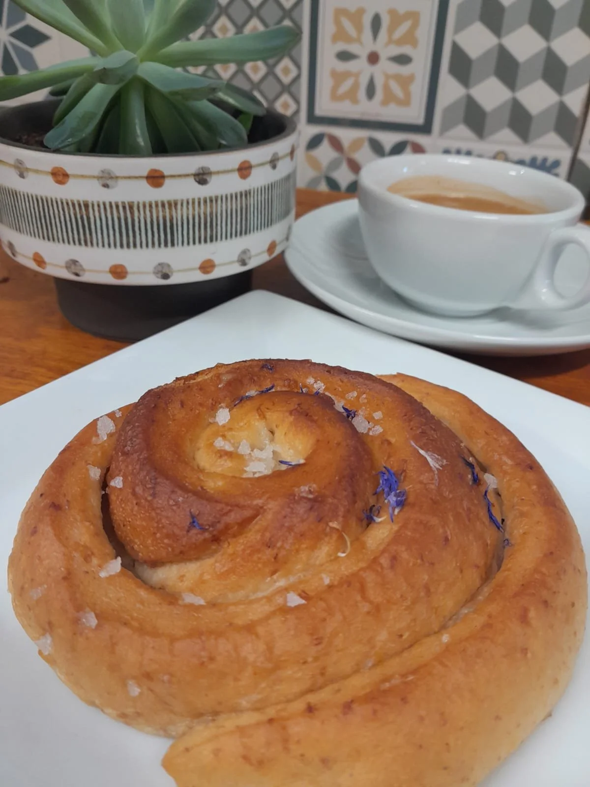 Close-up of a cinnamon roll garnished with salt flakes and purple edible flowers on a white plate. In the background, there is a cup of coffee or espresso in a white cup with a saucer, and a potted succulent plant with a patterned tile wall behind it