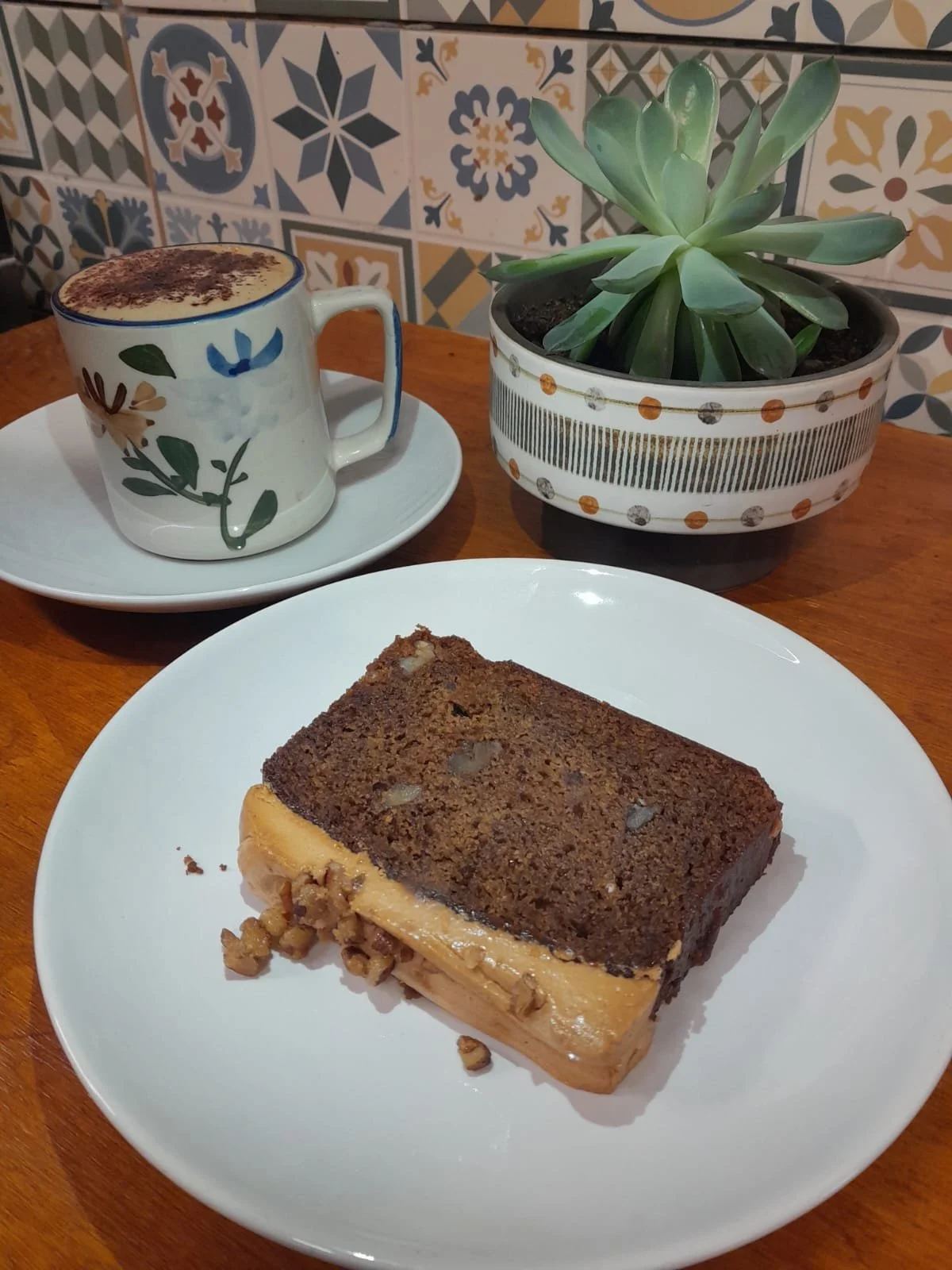 A slice of layered dessert on a white plate, a coffee mug with a floral pattern containing a coffee drink topped with cocoa powder, a potted succulent plant with thick, green leaves, and patterned tiles on the wall in the background.