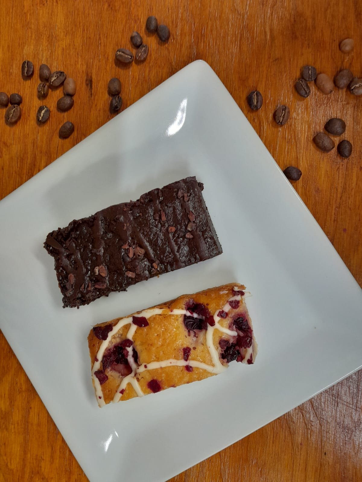 A white square plate with two slices of dessert on a wooden table. One slice is chocolate with chocolate shavings on top, and the other is a fruit-topped dessert with white drizzle and dried fruit pieces.
