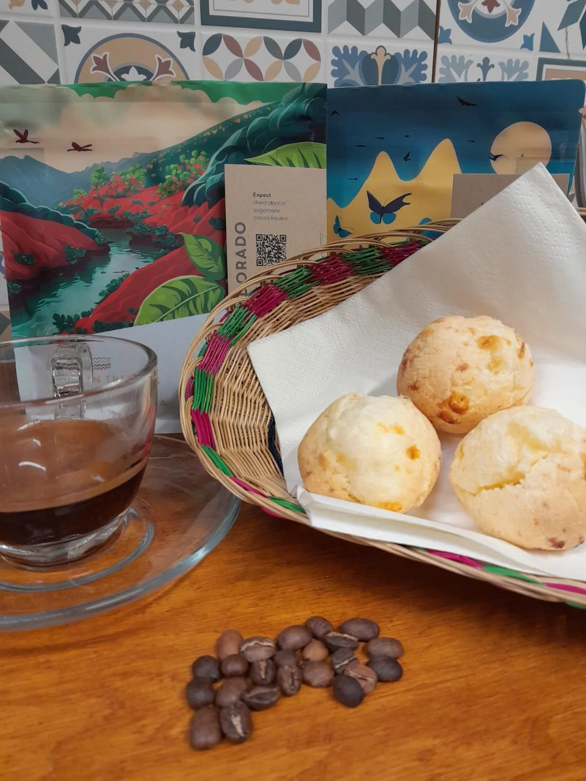 A basket with three cheese bread rolls lined with a white napkin, a glass cup with a dark coffee drink on a wooden table, a small pile of coffee beans, colorful patterned paper bags in the background, and a few wallpaper tiles with geometric and flor