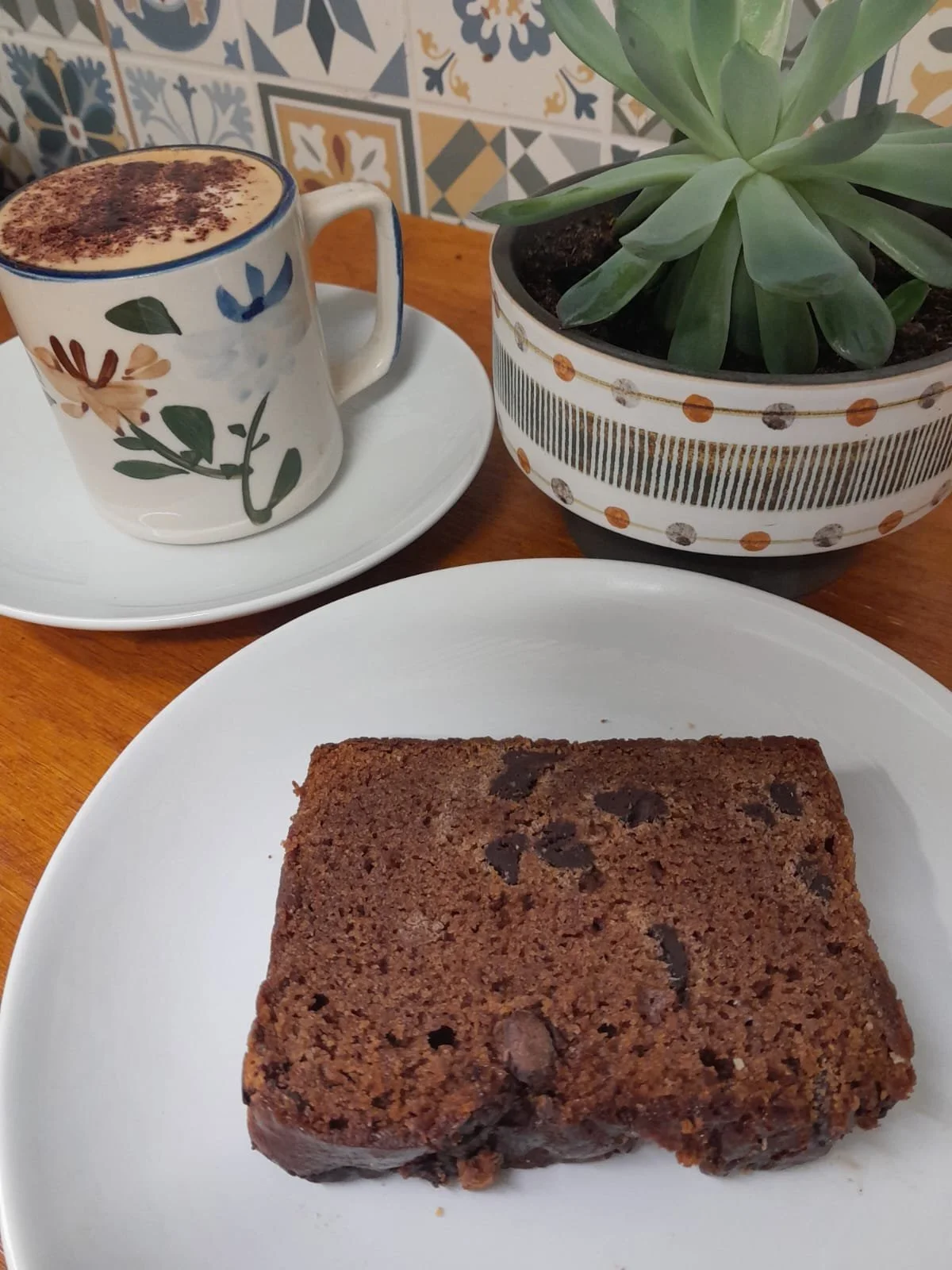 A square chocolate chip brownie on a white plate, a cup of coffee with foam and cocoa powder, a potted succulent plant, and a decorative tiled wall in the background.
