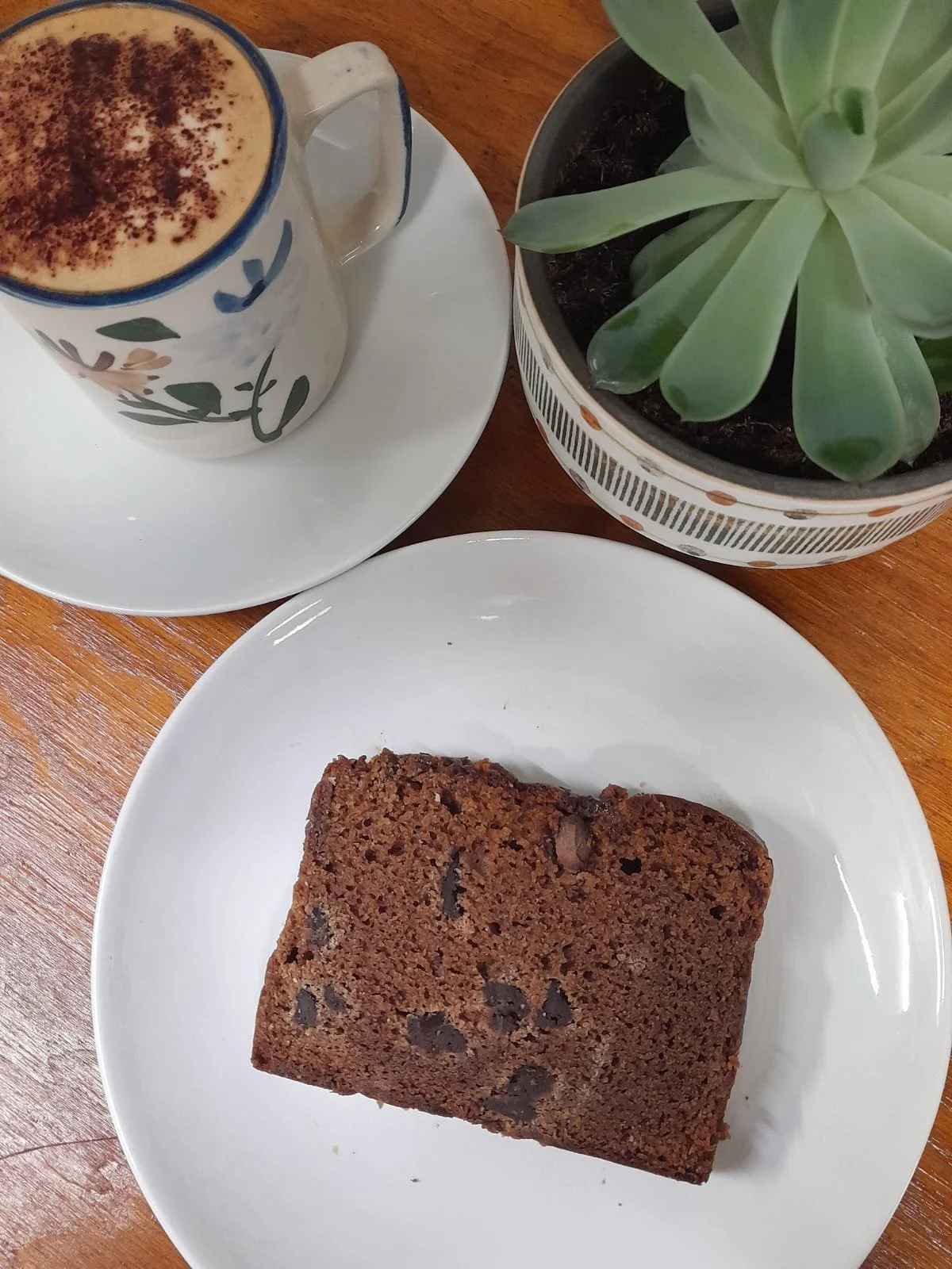 A slice of chocolate chip banana bread on a white plate with a cup of coffee topped with foam and cinnamon, and a potted succulent plant on a wooden table.