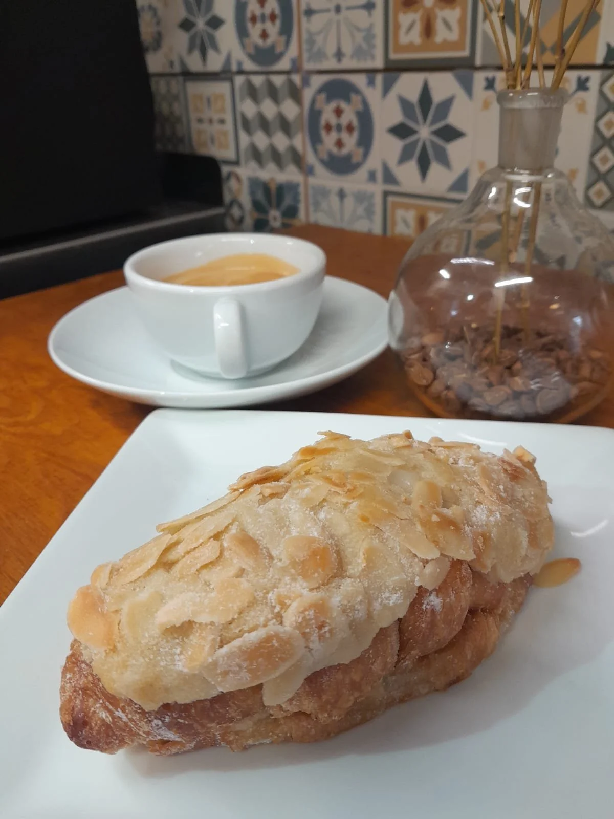A plate with an almond croissant on a white square plate, a cup of coffee on a saucer, and a glass jar with a few coffee beans and stirring sticks inside, set on a wooden table with a decorative tiled wall in the background.