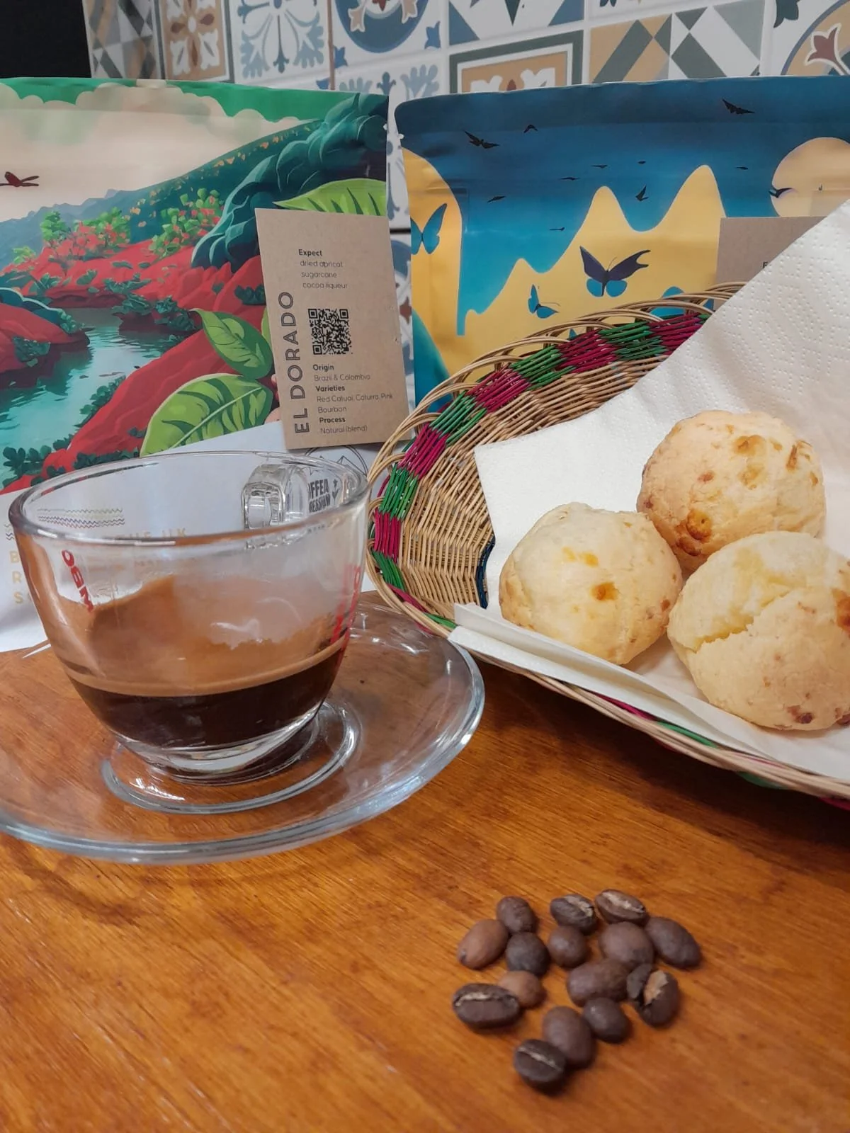 A cup of black coffee on a glass saucer, a basket with three bread rolls on a white napkin, a small pile of coffee beans on a wooden table, and colorful illustrated packages of coffee beans in the background.