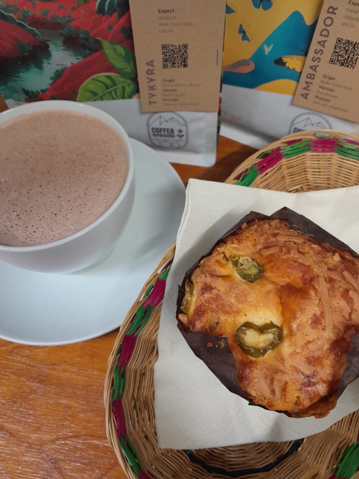 A cup of hot chocolate or coffee, a slice of savory bread or pizza with green peppers, and two packages of coffee beans on a wooden table.