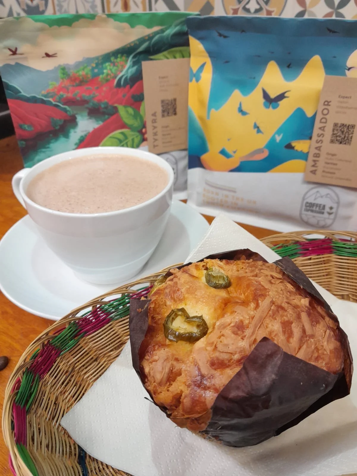 A basket containing a cheese bread with jalapeño slices, a white cup of hot chocolate on a saucer, and two bags of coffee beans in the background.