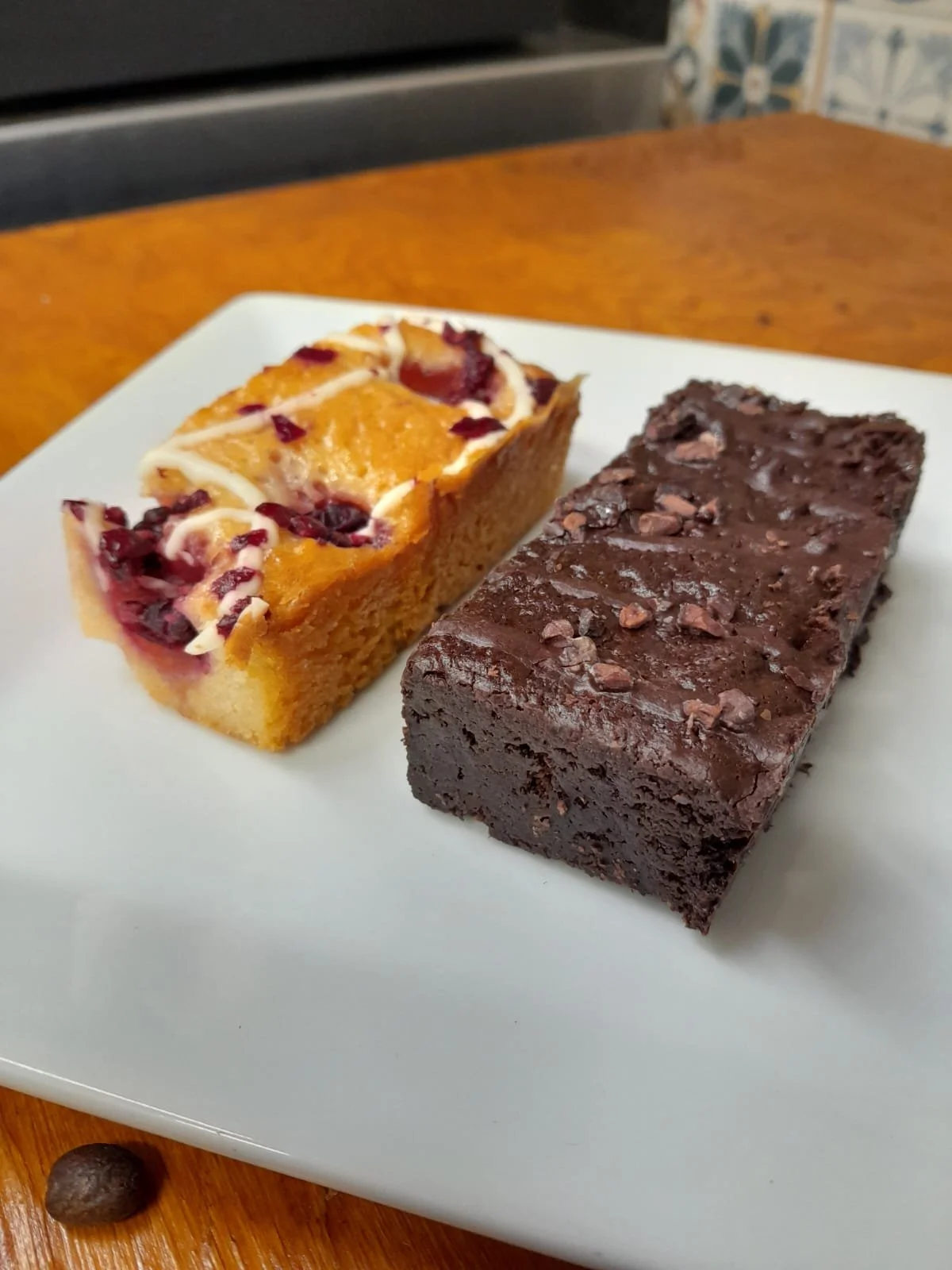 Two dessert bars on a white plate: a lemon bar with a crumbly crust, topped with white icing and dried cranberries, and a chocolate brownie with chocolate chunks and chopped nuts.
