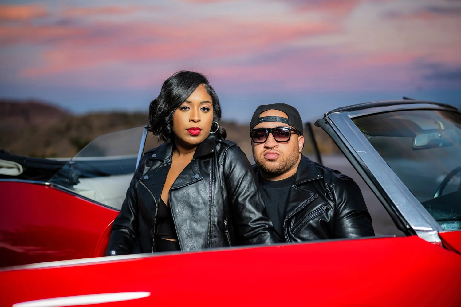 Two people, a woman and a man, sitting in a red convertible car during sunset. The woman has short black hair, red lipstick, and wears a black leather jacket. The man has a beard, sunglasses, a black cap backwards, and a black leather jacket.