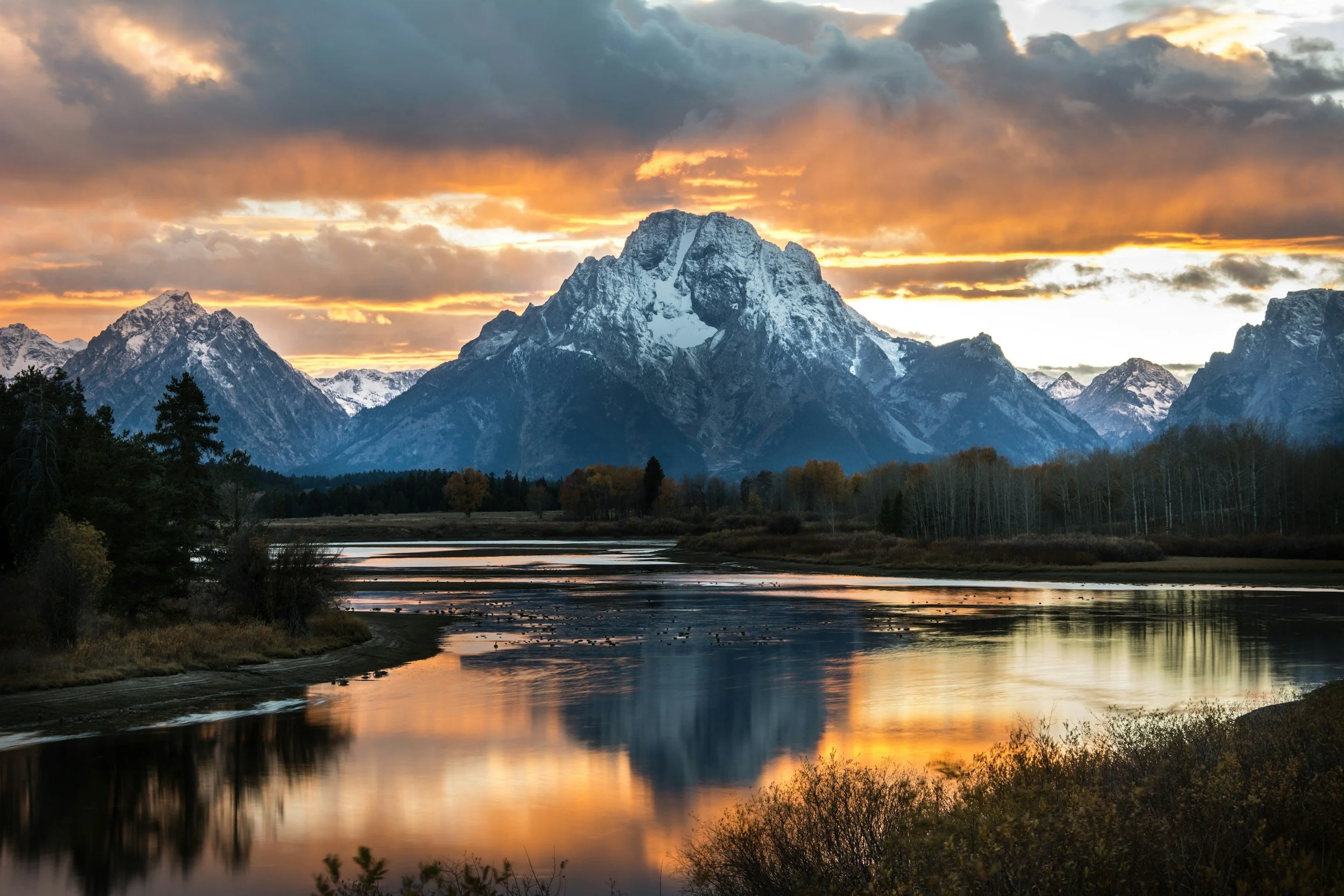Snow-capped mountain range at sunset with dark clouds and orange hues, reflection on a calm river in the foreground, surrounded by forested trees.