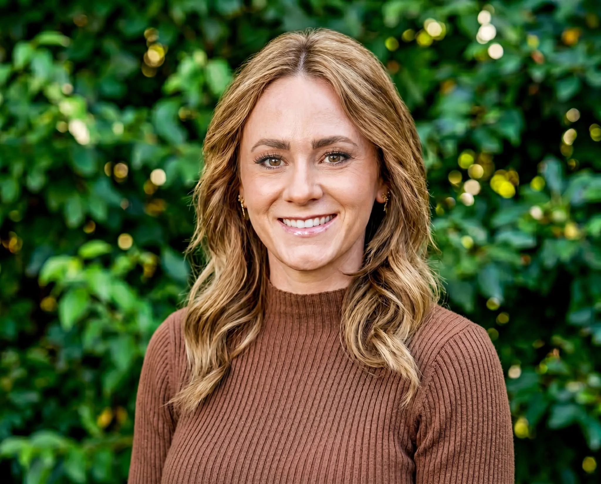 Alec has with light brown, wavy hair and brown eyes smiling in front of a green leafy background, wearing a brown ribbed top.