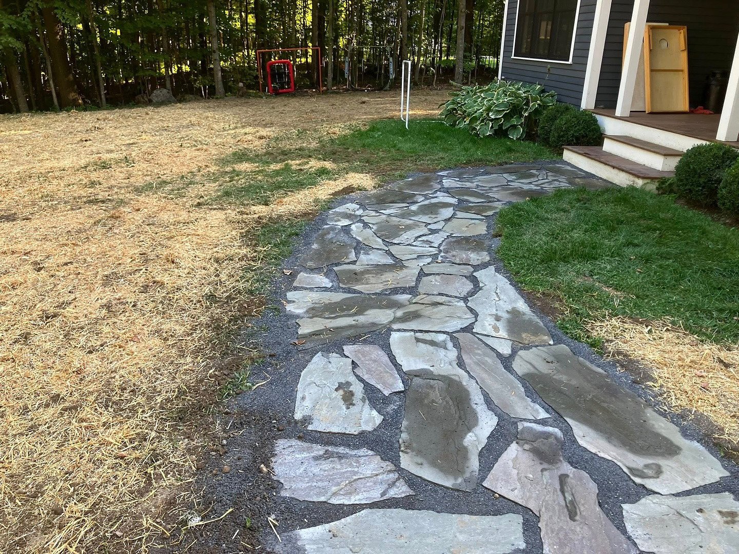 A stone pathway leading from a house porch into a grassy backyard with trees in the background, some bushes along the house, and construction tools and materials on the porch.