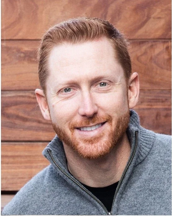 A professional headshot of a man in a dark suit, white shirt, and dark tie, smiling against a plain gray background.