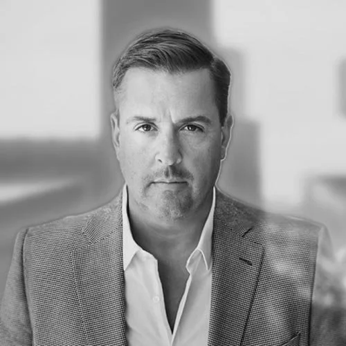 A professional headshot of a man in a dark suit, white shirt, and dark tie, smiling against a plain gray background.