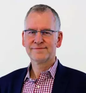 A middle-aged man with short gray hair, glasses, wearing a dark blazer and a checkered shirt, standing against a plain white background.