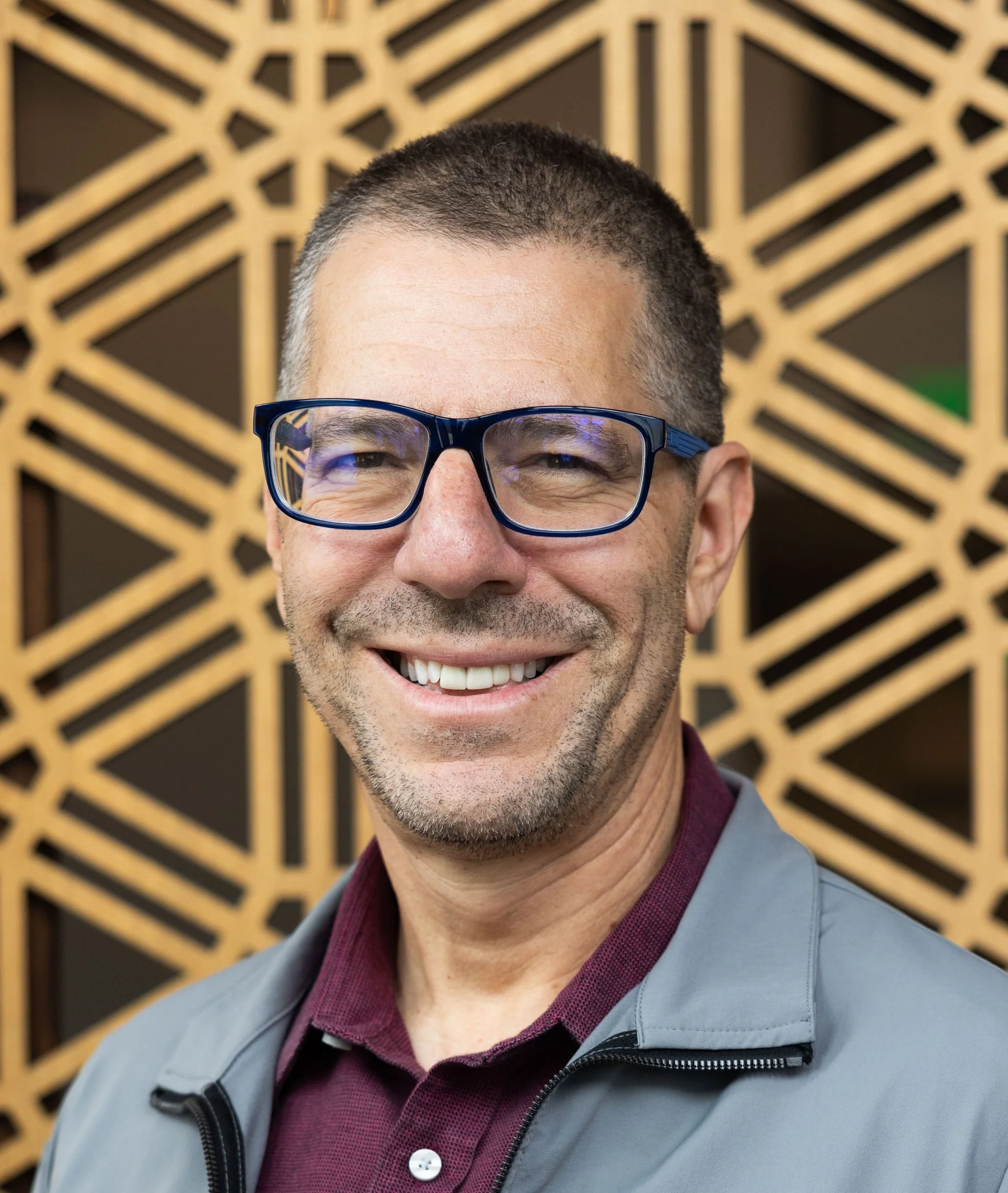 A man with short dark hair, glasses, and a beard wearing a navy blue shirt, smiling at the camera against a plain light-colored background.