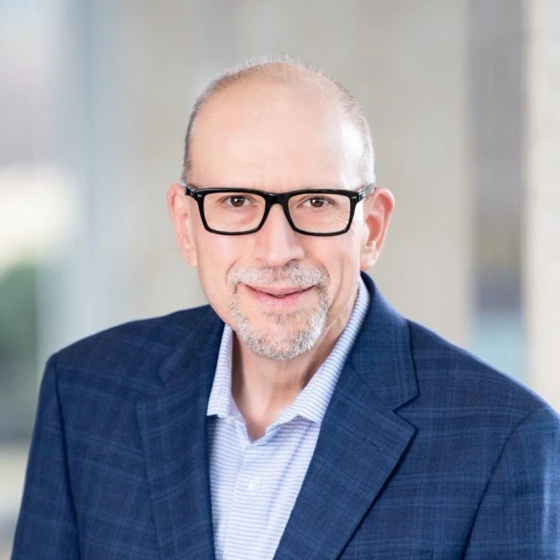 A middle-aged man with a bald head, glasses, and a beard, wearing a blue suit jacket and a light blue dress shirt, smiling in a professional indoor setting.