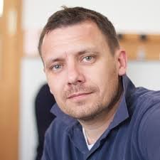 A man with short brown hair and light skin taking a selfie indoors, wearing a blue shirt.