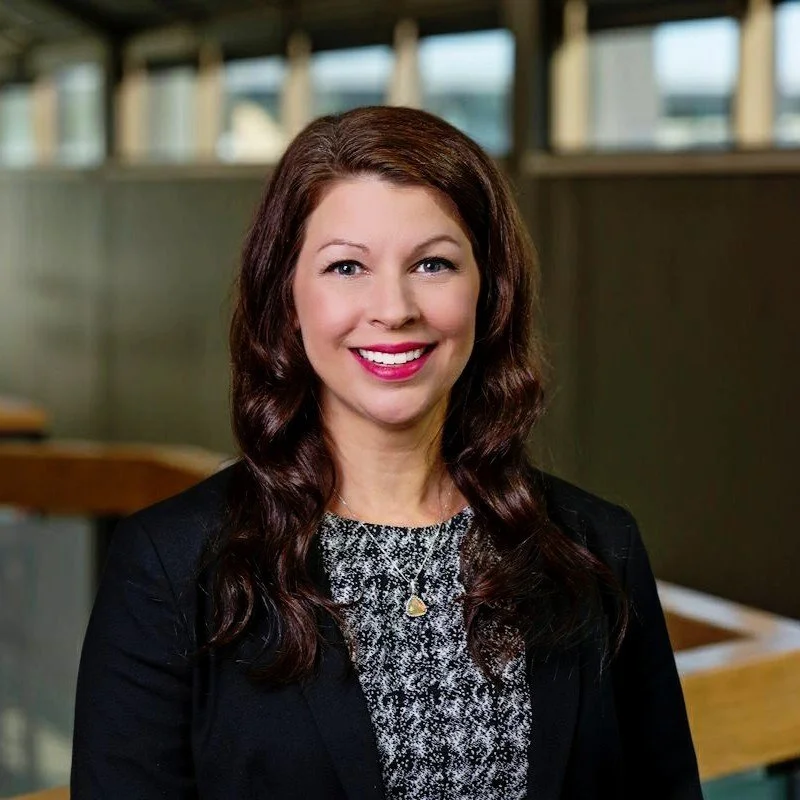 A smiling woman with long, wavy brown hair, wearing a black blazer, patterned top, and a gold heart necklace, standing indoors with blurred windows in the background.