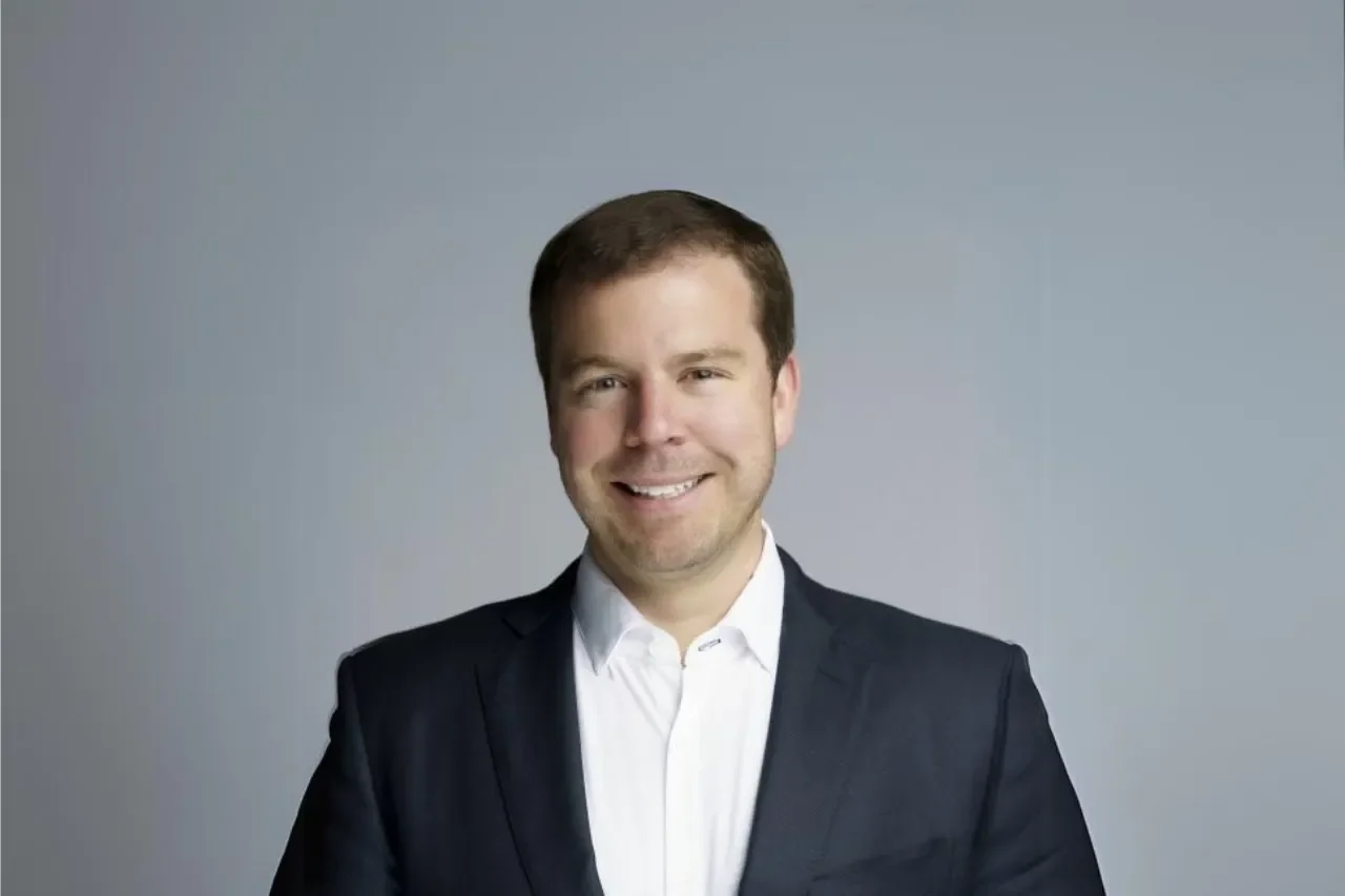 A smiling woman with shoulder-length blonde hair, wearing a yellow top, outdoors with a blurred background of a lawn and building.