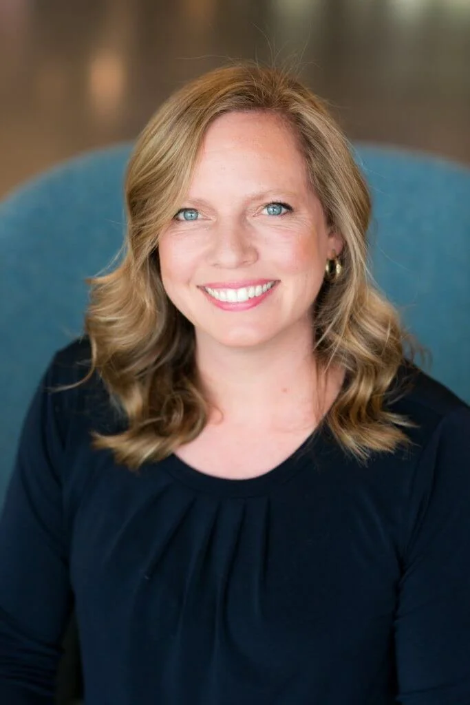 Portrait of a young woman with long brown hair, wearing a light blue top, standing against a gray background.