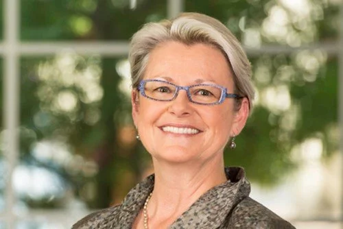 A smiling middle-aged woman with short gray hair, wearing glasses and a patterned blazer, outdoors with blurred greenery in the background.