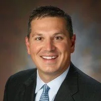Portrait of a young woman with long brown hair, wearing a light blue top, standing against a gray background.