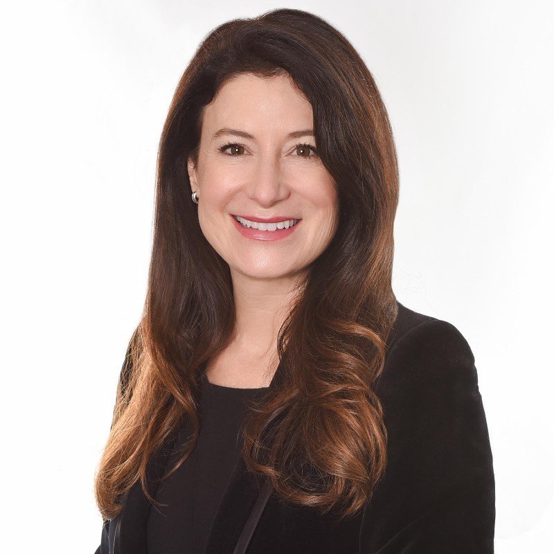 Portrait of a woman with long brown hair, wearing a black blazer and smiling at the camera.