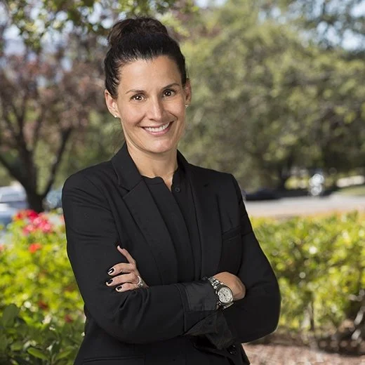 A woman with shoulder-length blonde hair smiling outdoors with a building in the background.