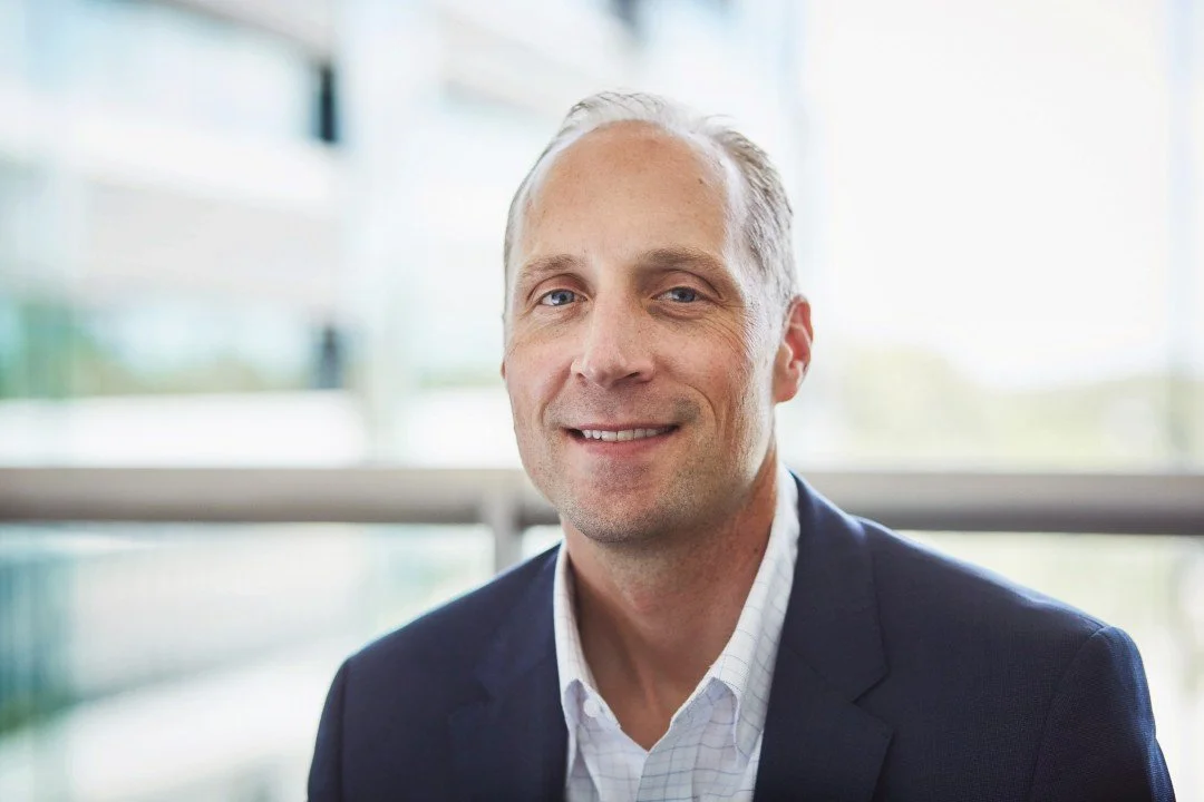 A middle-aged man with short, light-colored hair, wearing a navy blazer and white shirt, smiling indoors with a bright, modern background.