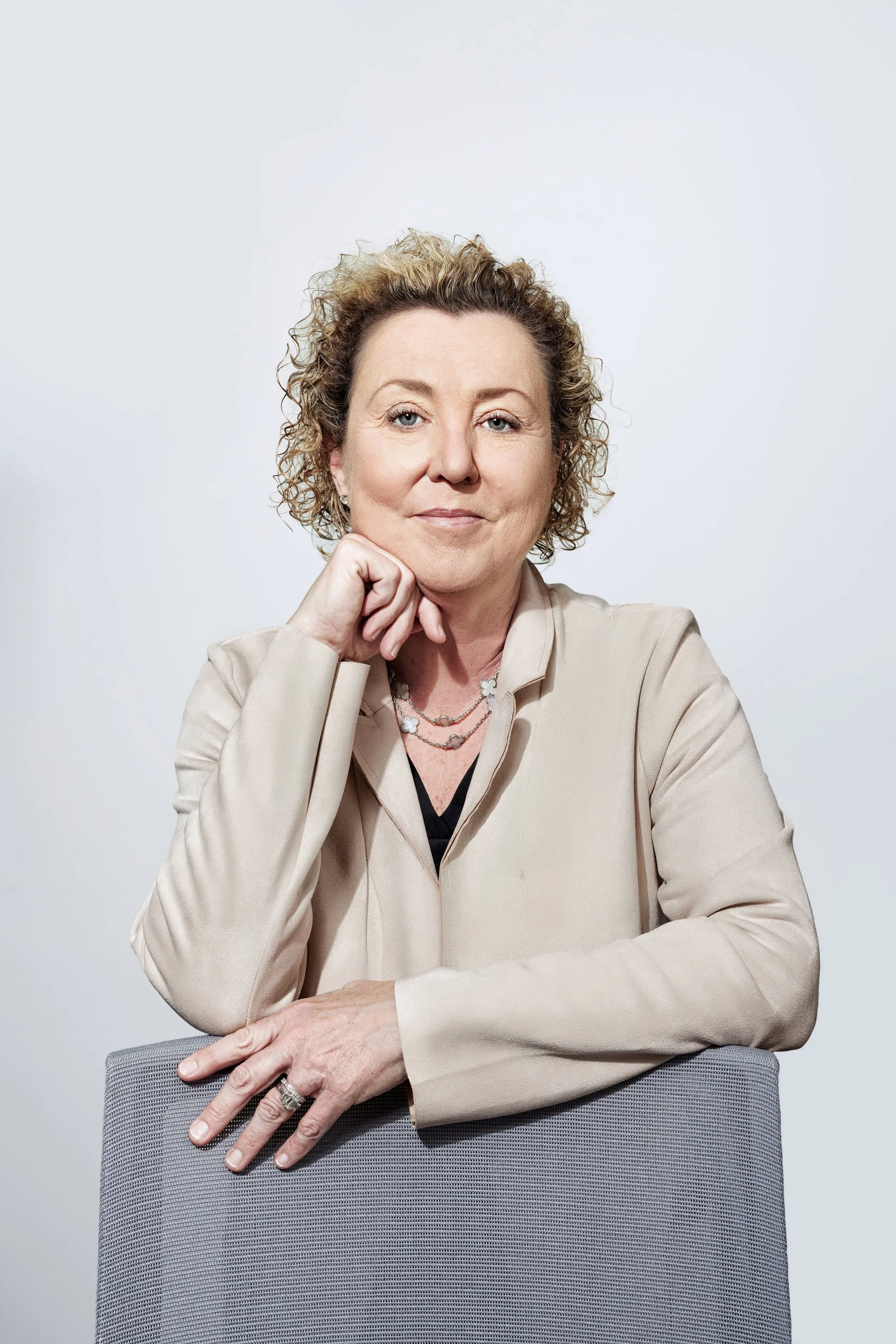 A woman with shoulder-length dark hair wearing a black top and layered pearl necklaces, standing in front of a plain gray background.