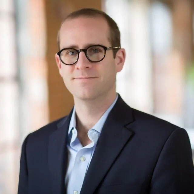 Professional headshot of a man wearing glasses, a navy blazer, and a light blue shirt in an indoor setting with windows in the background.