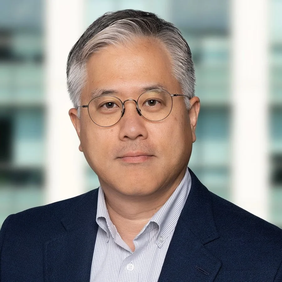 A professional headshot of a middle-aged Asian man with gray hair and glasses, wearing a dark blazer and a collared shirt, with a blurred cityscape background.