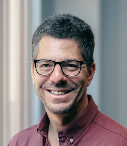 Close-up of a smiling man with glasses, dark hair with gray, wearing a maroon shirt, indoors near a window.