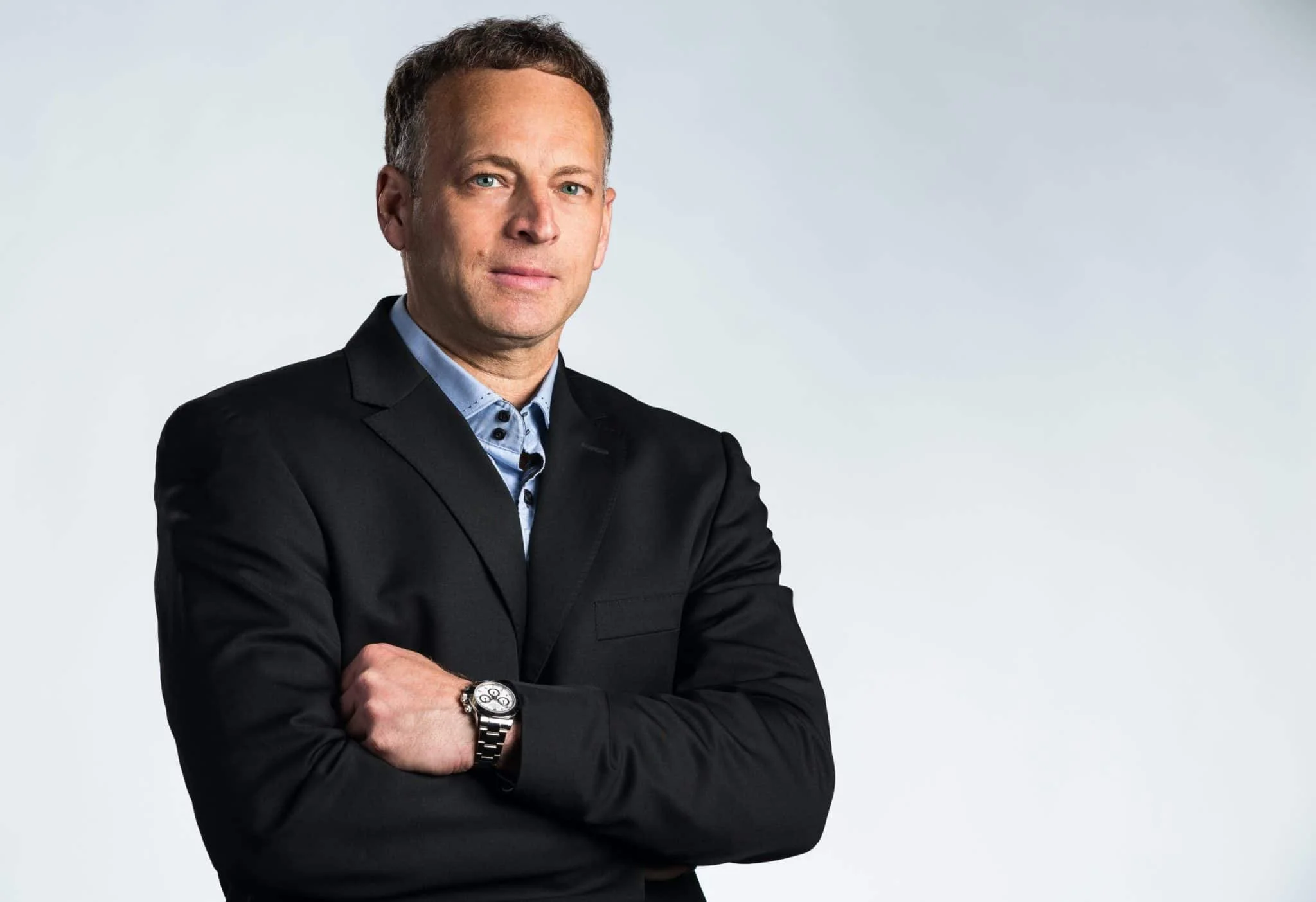A man wearing a black blazer and a watch, standing with arms crossed against a plain light gray background.