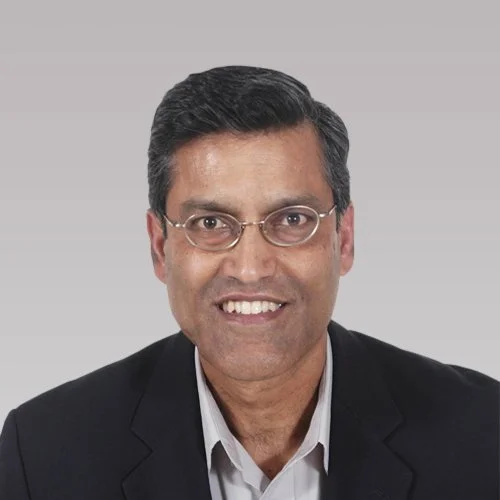 Portrait of a young woman with long brown hair, wearing a light blue top, standing against a gray background.