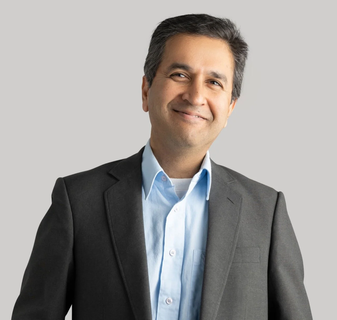 Professional middle-aged man in a gray suit and light blue shirt, smiling, standing against a plain light gray background.