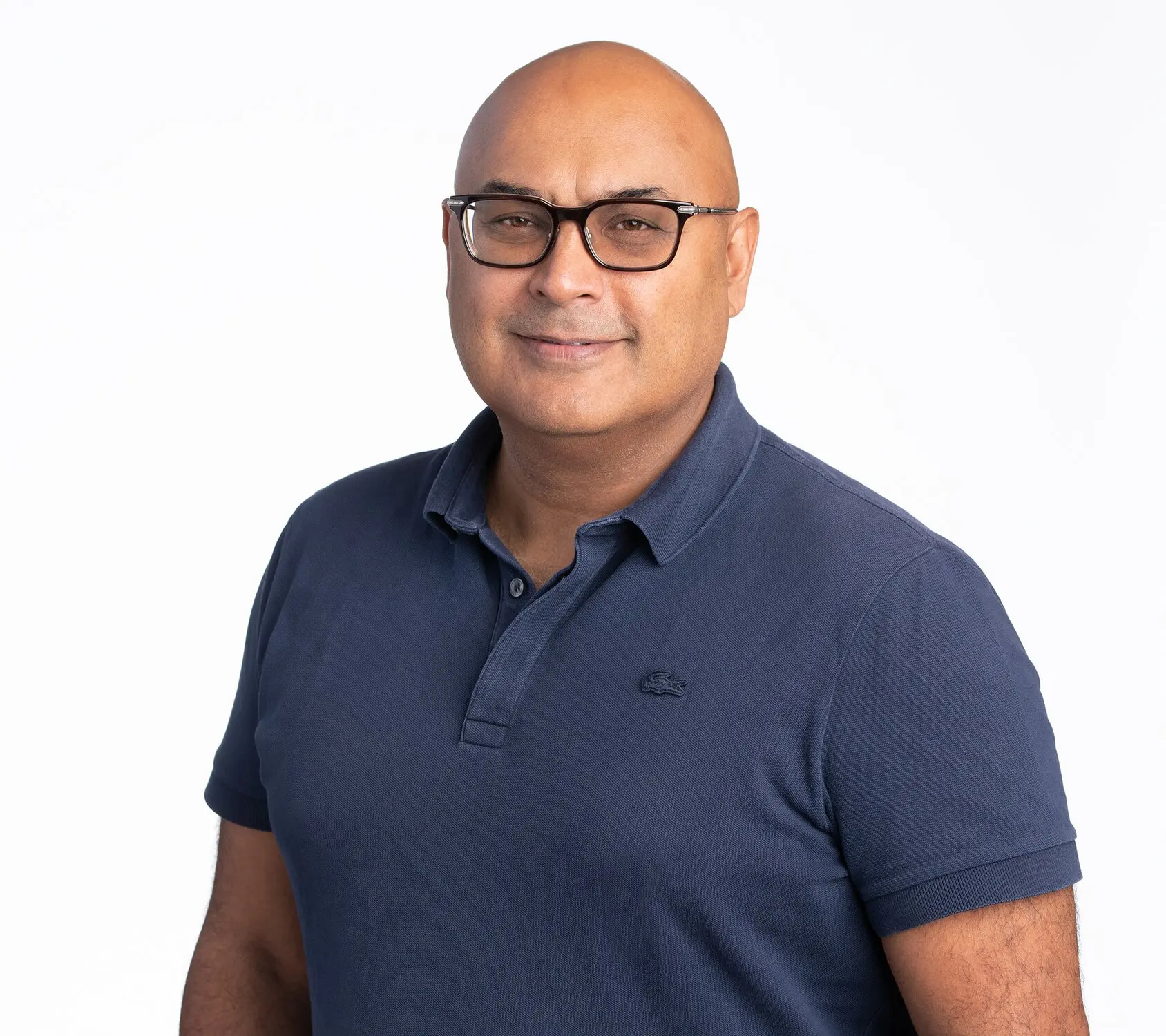 A smiling man with short hair, wearing a white shirt and a blue plaid blazer, against a dark background.