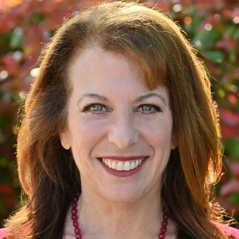 Portrait of a young woman with long brown hair, wearing a light blue top, standing against a gray background.
