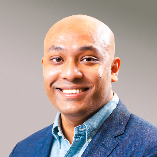 Close-up of a smiling man with a shaved head, wearing a blue blazer and light blue collared shirt, against a neutral background.