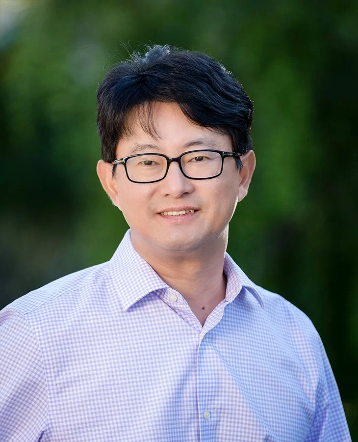 Headshot of a man with black hair, glasses, wearing a light purple checkered shirt, smiling outdoors with greenery in the background.