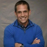 Professional man wearing a dark suit and light blue checkered shirt, smiling at the camera against a gray background.