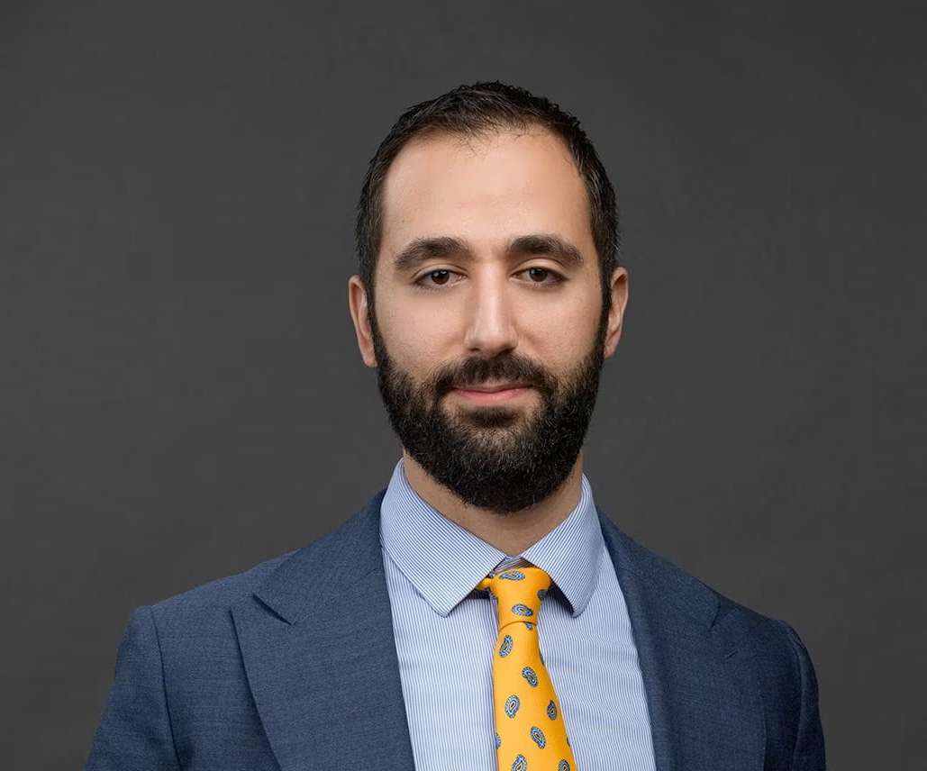Professional headshot of a man with dark hair and a beard, wearing a blue suit, light blue shirt, and yellow tie with blue patterns, standing against a dark grey background.