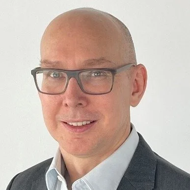 A woman with shoulder-length blonde hair smiling outdoors with a building in the background.