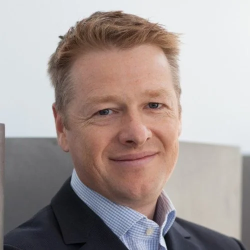 Close-up of a smiling man in a suit and collared shirt, with a blurred outdoor background.